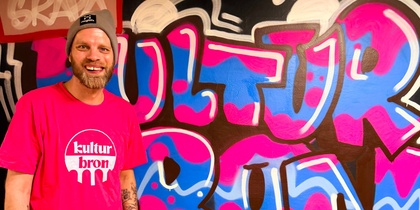 Person i rosa t-shirt med texten 'kultur bron' står framför färgglad graffiti-vägg.