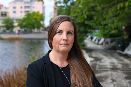 Kvinna med långt brunt hår står utomhus vid en vattendrag och grönska, bär svart kavaj.