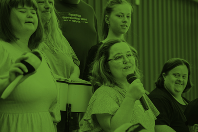 En grupp människor, varav en kvinna håller en mikrofon, sitter tillsammans inomhus. Bilden har grön färgton.