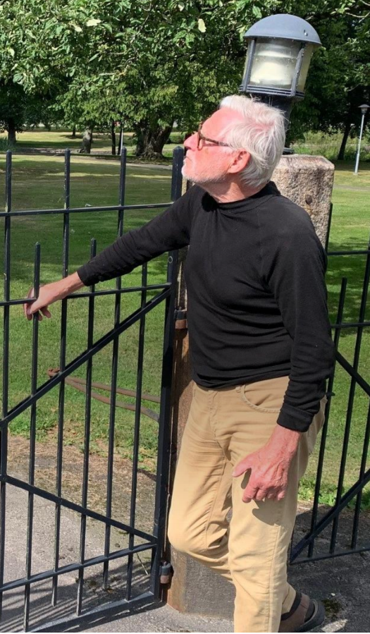 En äldre man med vitt hår och glasögon står vid en svart järngrind i en park, klädd i svart tröja och beige byxor.