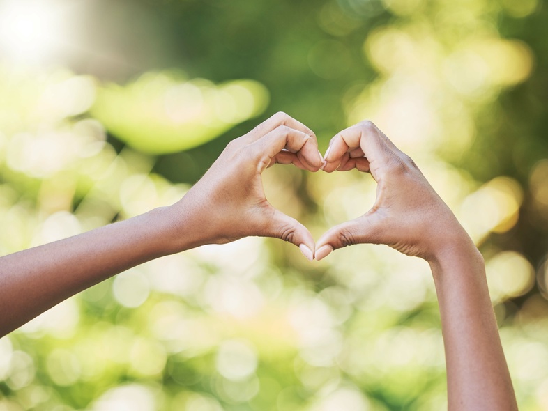 Två händer formar ett hjärta mot en suddig, grön bakgrund utomhus.