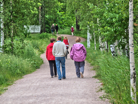 Flera personer promenerar på en grusväg omgiven av gröna träd och gräs en sommardag.
