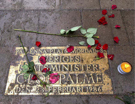 Minnesplakett på trottoar med rosor, rosenblad och ett tänt ljus till minne av Olof Palme.