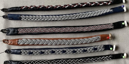 Sex handgjorda armband i läder och tenntråd med olika flätade mönster, liggande på ett grått tyg.