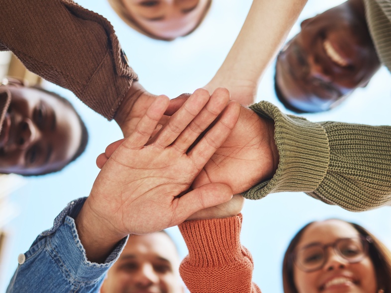 Fem personer står i en cirkel och lägger sina händer ovanpå varandra, sedda underifrån mot himlen.