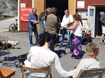 En grupp människor samlade utomhus vid en röd byggnad, några sitter på stolar och andra står och pratar.