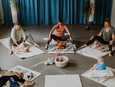 Fyra vuxna och tre bebisar sitter på mattor i en cirkel i ett ljust rum med blå gardiner.