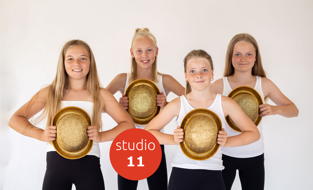 Fyra unga tjejer i vita linnen och svarta byxor håller gyllene hattar framför sig, står mot vit bakgrund.