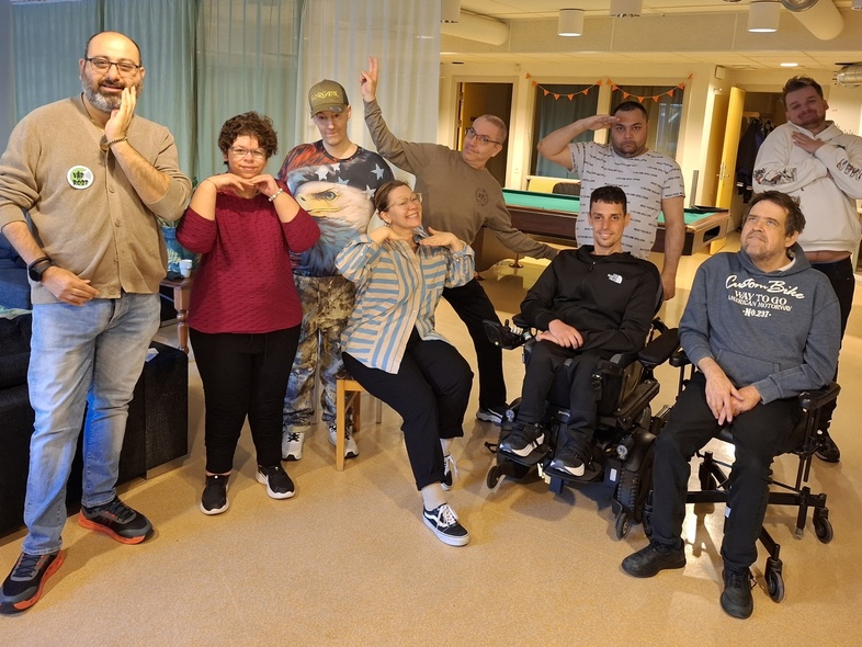 En grupp människor poserar glatt tillsammans inomhus, några sitter i rullstol, andra står eller gör roliga poser.