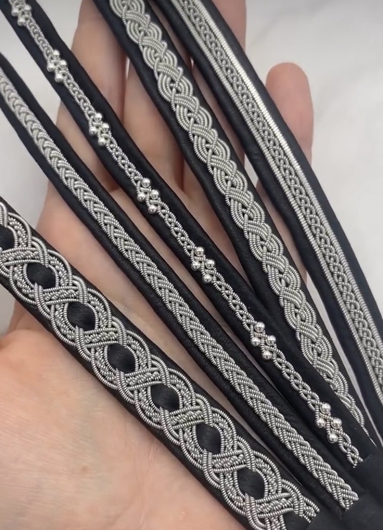 Hand håller flera svarta läderband med intrikata silverbroderier i olika mönster.