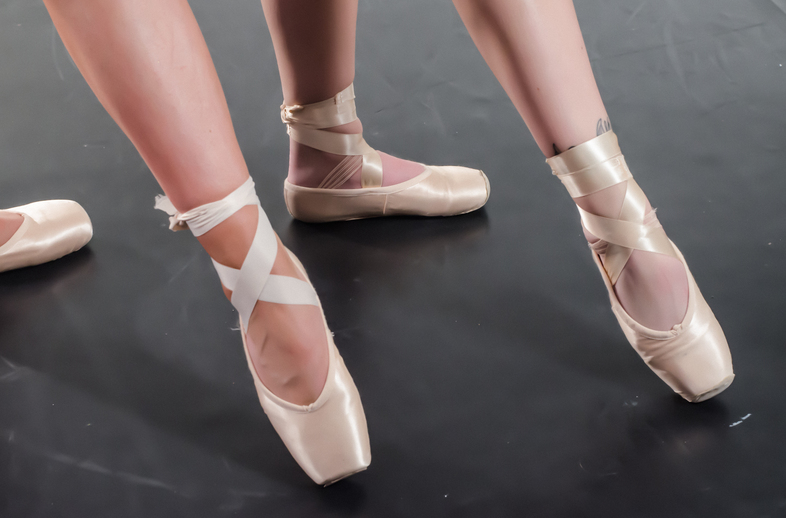 Två personer står på tå i balettskor på ett mörkt golv, endast benen och fötterna syns.