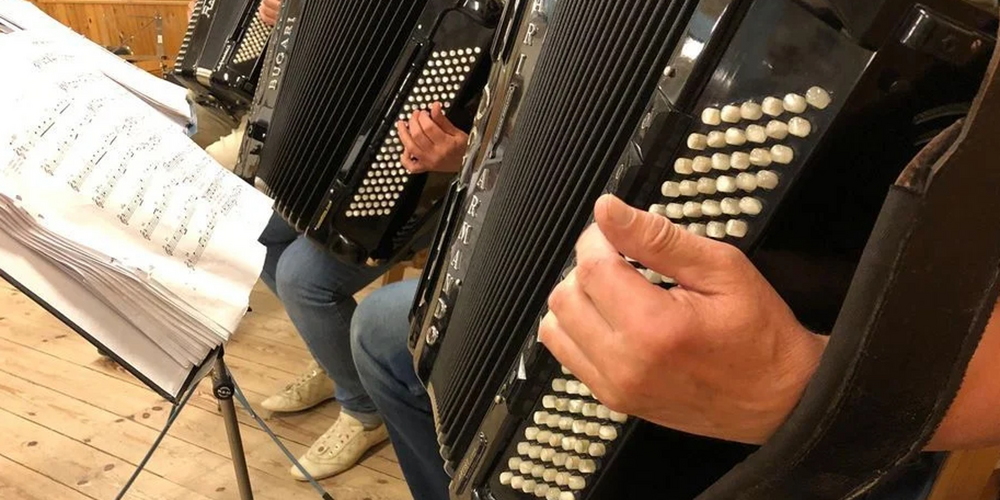 Två personer spelar dragspel vid ett notställ med noter i ett trägolv-rum.