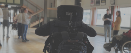 Person i rullstol i förgrunden, flera personer och en annan rullstolsburen i bakgrunden i en ljus sal.