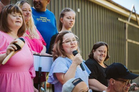 En grupp människor, flera med mikrofon och musikinstrument, står utomhus och sjunger tillsammans.