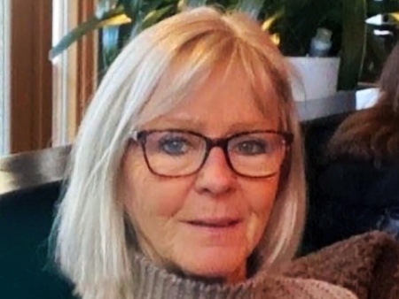 Kvinna med blont hår och glasögon sitter inomhus i en mysig miljö med växter i bakgrunden.