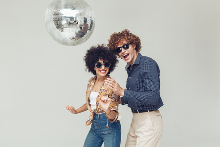 Två personer med lockigt hår och solglasögon dansar under en discokula mot en ljus bakgrund.
