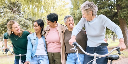 Fem kvinnor i olika åldrar skrattar tillsammans utomhus med cyklar och skateboard.