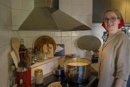 Kvinna med glasögon och sjalett lagar mat i ett kök och ler mot kameran.