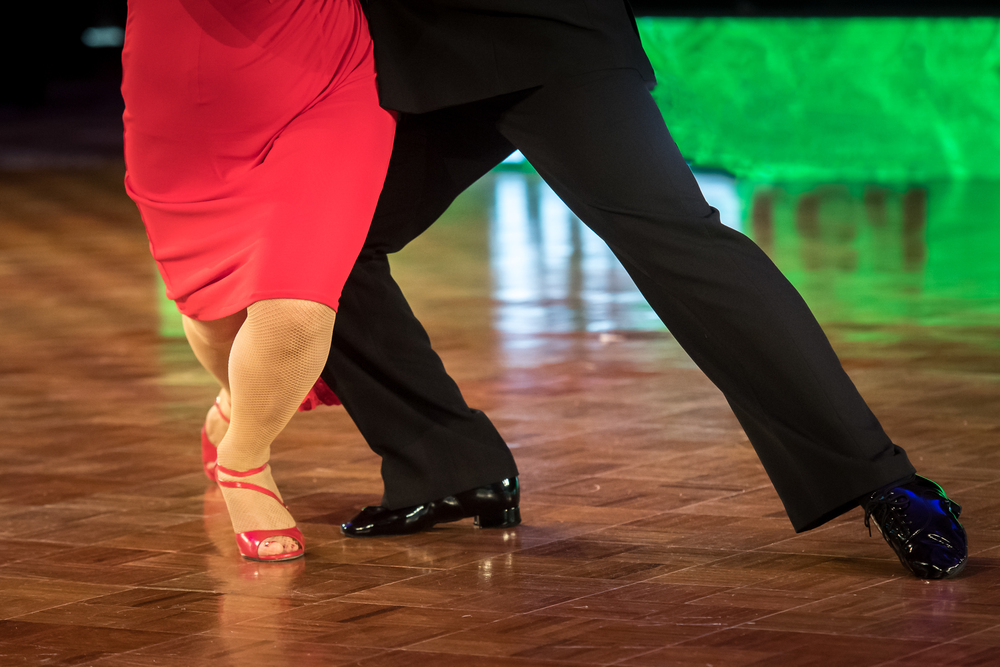Två personer dansar tillsammans på ett dansgolv, endast deras ben och fötter syns.