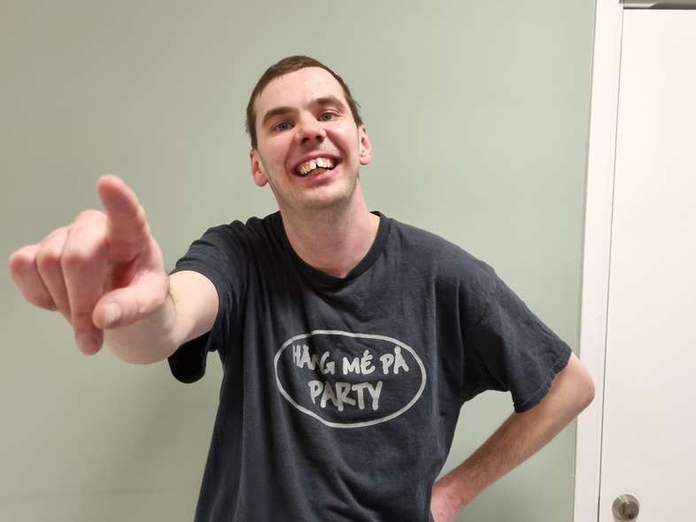 Person i svart t-shirt pekar mot kameran, ansiktet är suddat, står vid en ljusgrön vägg och vit dörr.