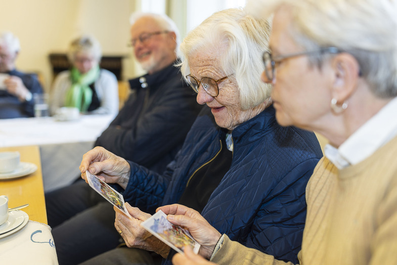 Två äldre kvinnor sitter vid ett bord och tittar på fotografier tillsammans, andra äldre personer i bakgrunden.