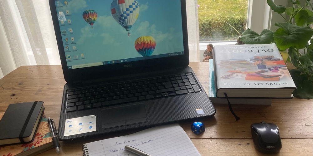 Ett skrivbord med laptop, anteckningsbok, penna, böcker, mus och växt vid fönster med utsikt mot gräsmatta.