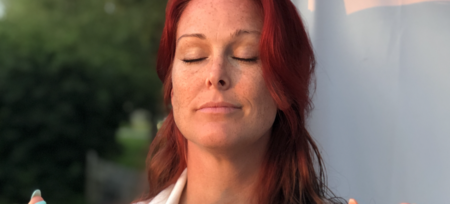 Kvinna med rött hår står utomhus med slutna ögon och ser avslappnad ut i mjukt ljus.