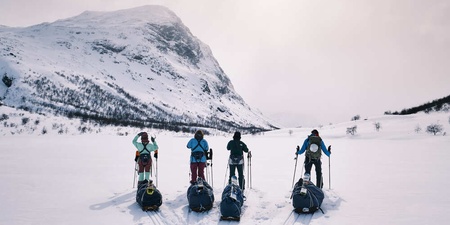 Fem personer med ryggsäckar och pulkor står på skidor i ett snötäckt fjällandskap med berg i bakgrunden.