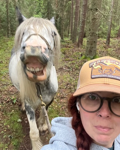 En kvinna med glasögon och keps tar en selfie med en häst som gör en rolig min i en skog.