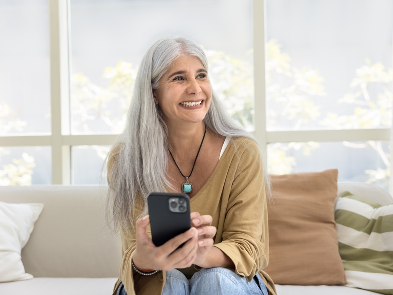 Person med långt grått hår sitter i soffa och håller en mobiltelefon i vardagsrum med stora fönster.