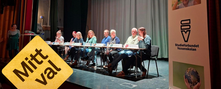 Panel med åtta personer sitter på scen, en person står bredvid. Skylt med texten 'Mitt val' och roll-up från Studieförbundet Vuxenskolan.