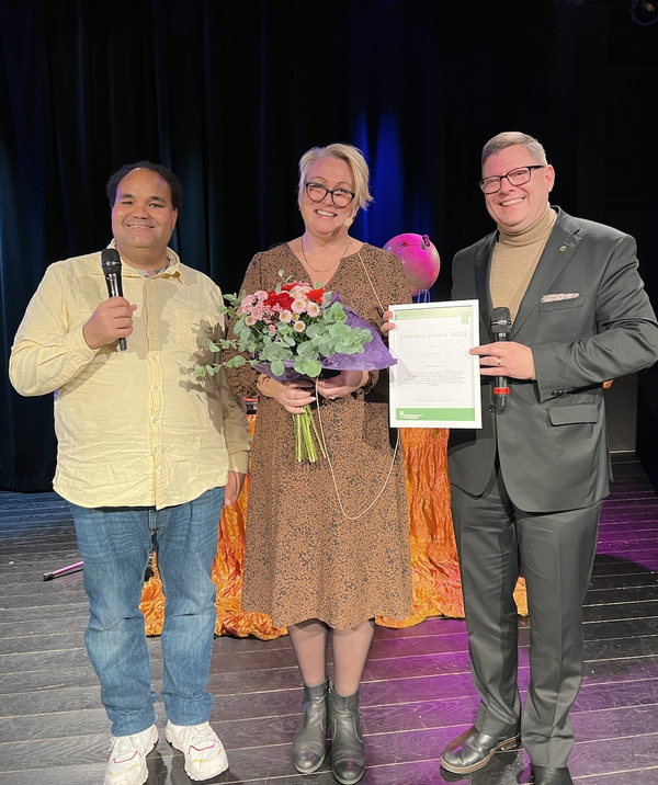 Tre personer står på en scen, en håller blommor och en annan ett diplom. Två har mikrofoner.