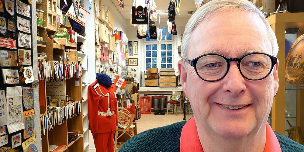 Äldre man med glasögon ler i ett rum fullt av samlarföremål och memorabilia.