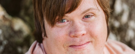 Ung man med ljust hår och rosa tröja sitter utomhus framför ett träd och ler mot kameran.