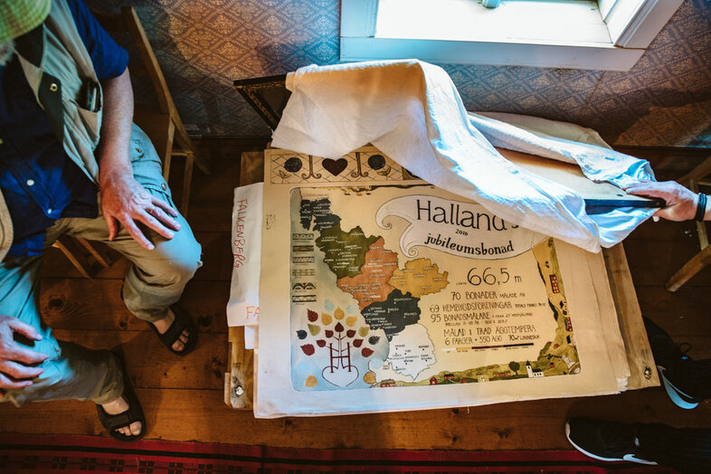Två personer sitter vid ett bord med en karta och text om Halland, delvis täckt av en duk.