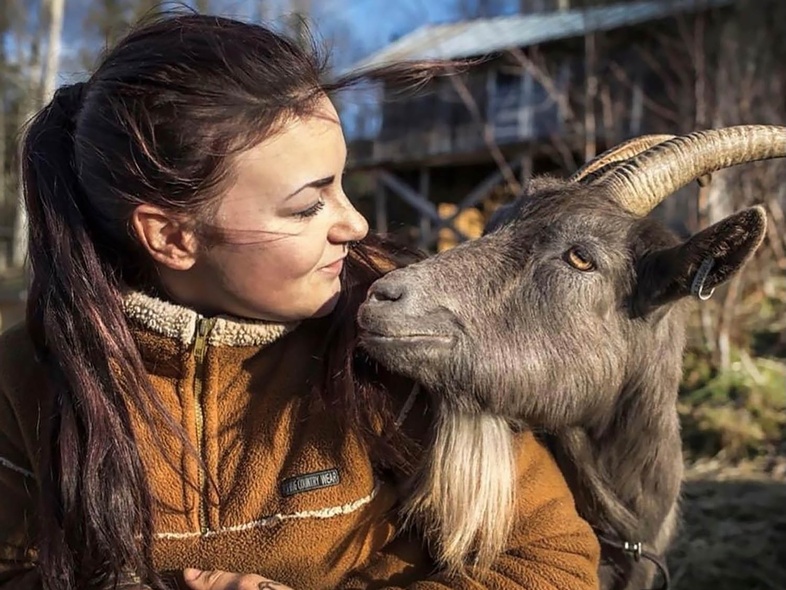 En kvinna i brun fleecejacka tittar leende på en get som står nära henne utomhus.