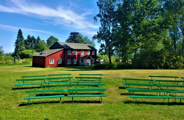 En lantlig bild med ett rött hus med vita knutar, en dunge träd och framför dessa står en några gröna bänkar utställda på gräset.