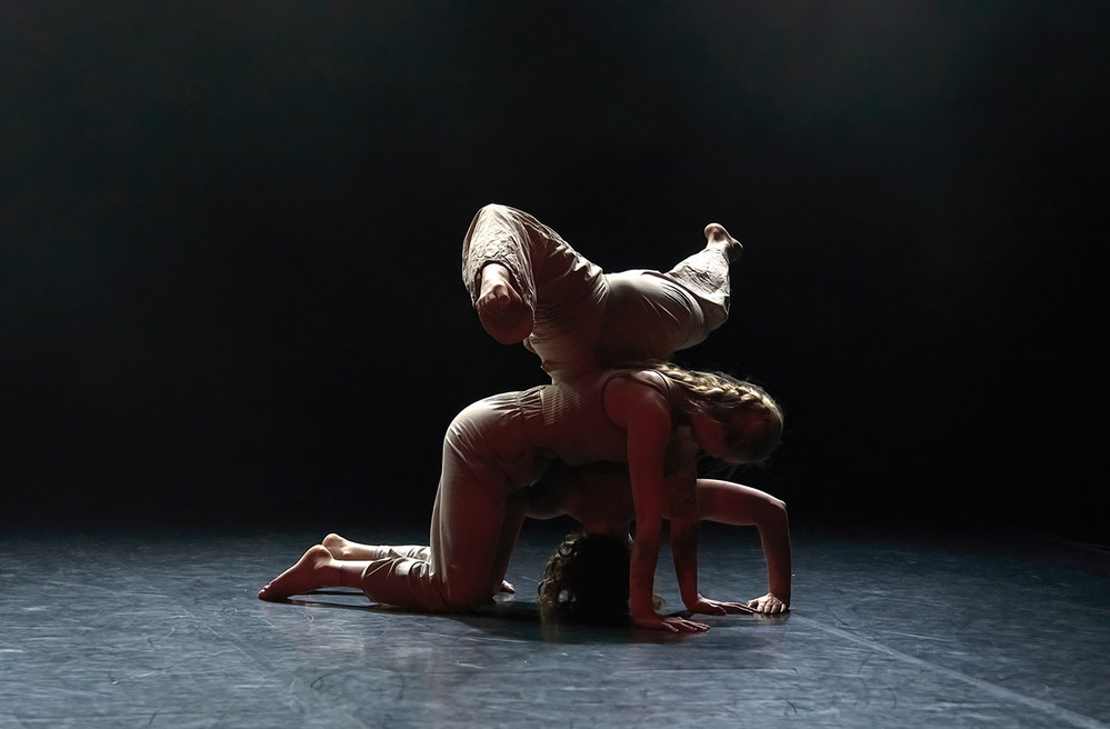 Två dansare utför en akrobatisk pose på en mörk scen, med en person balanserande ovanpå den andra.