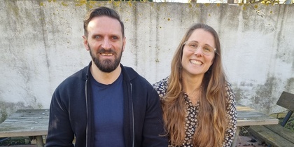 Två personer sitter på en bänk utomhus med en svart ledarhund mellan sig.