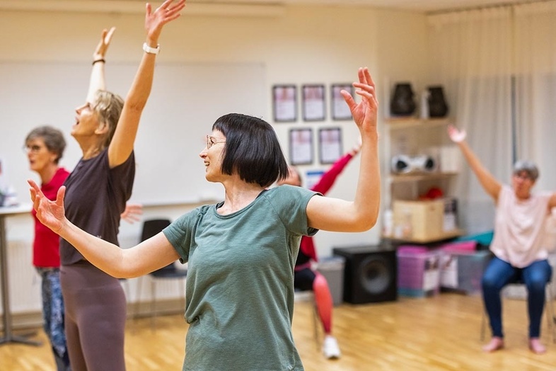 En grupp vuxna deltar i en dansklass i en ljus träningssal, med armarna uppsträckta och leenden på läpparna.