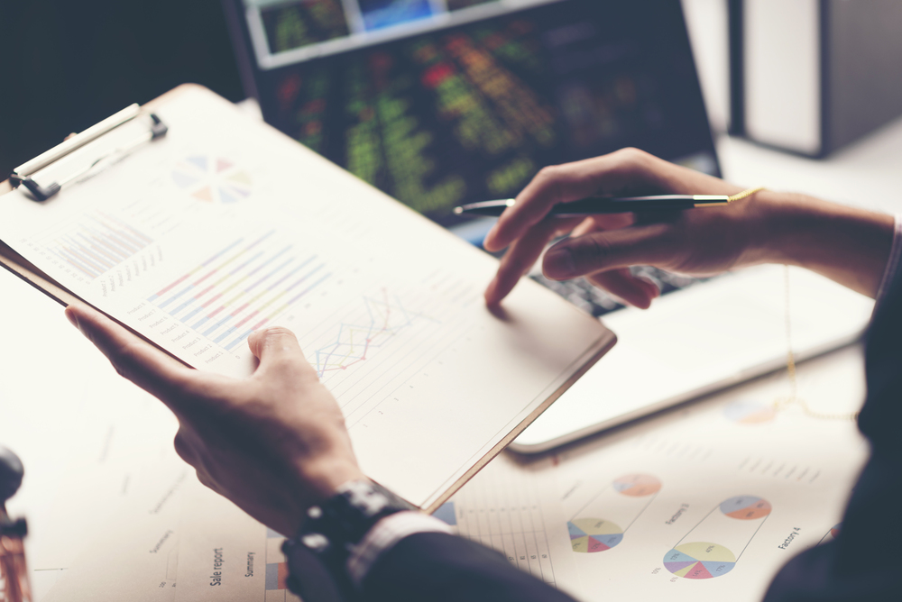 En person granskar diagram och grafer på ett papper med en penna, med en bärbar dator i bakgrunden.