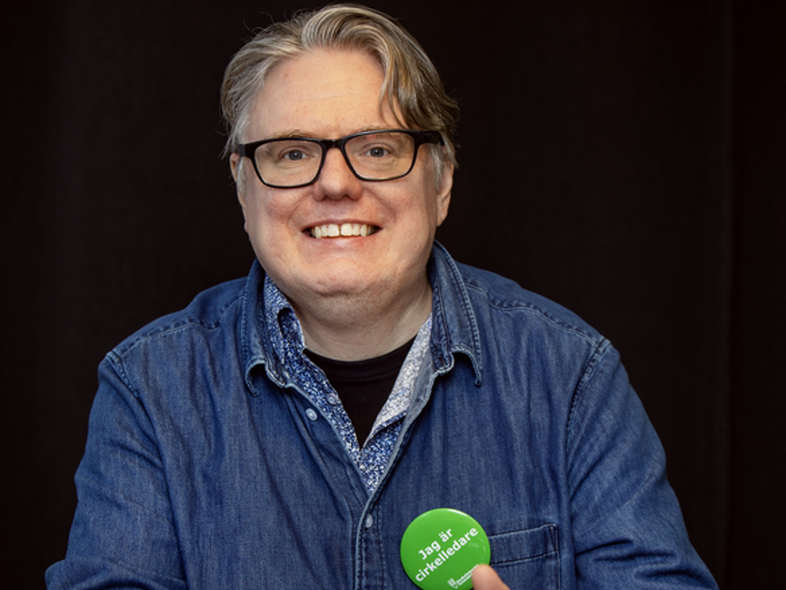 Leende person med glasögon och jeansskjorta sitter vid ett bord och håller en grön knapp med text.