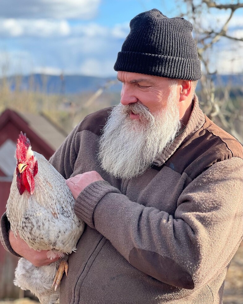 En äldre man med grått skägg och svart mössa håller en höna i famnen utomhus.