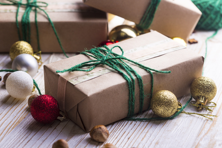 Inslagna paket med brunt papper och grönt snöre, omgivna av julkulor och nötter på ett trägolv.