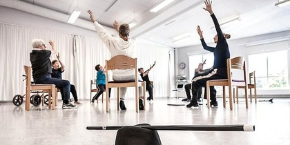 En grupp personer sitter på stolar i en ljus sal och sträcker armarna uppåt, hatt och käpp i förgrunden.