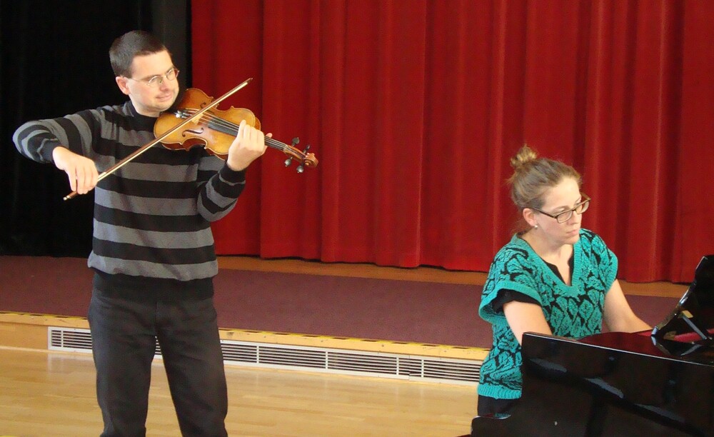 En person spelar fiol och en annan person spelar piano på en scen med röd ridå i bakgrunden.