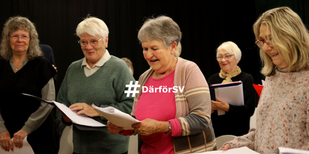 En grupp äldre personer står tillsammans och sjunger, håller papper och ser glada ut.