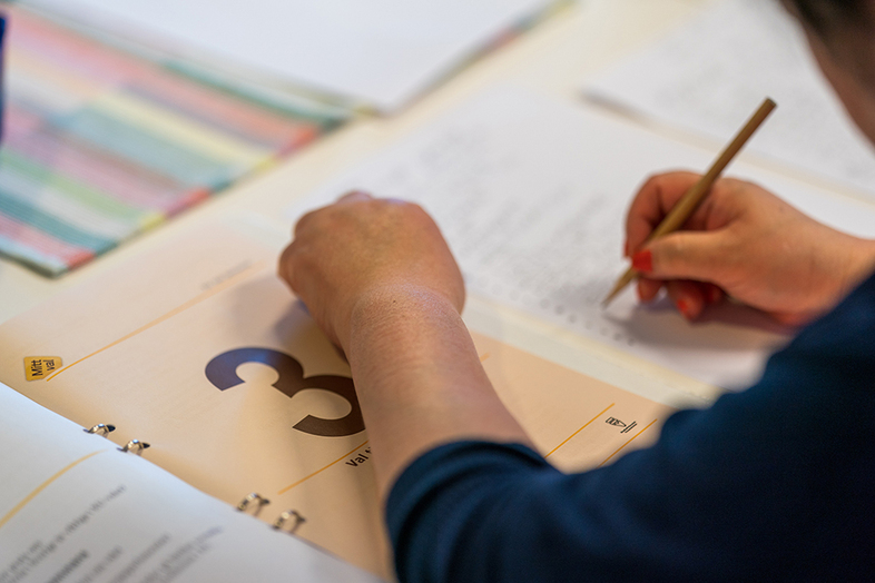 En person skriver med penna på papper bredvid en bok med siffran 3 på omslaget.