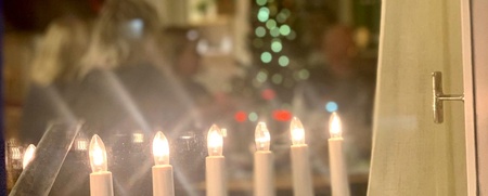 Ett fönster med elektriska ljusstakar och suddig julgran i bakgrunden, människor sitter inomhus.