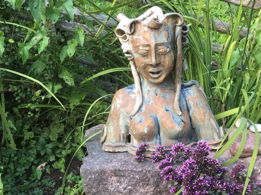 Keramikskulptur av en kvinna med långt hår står på en sten omgiven av gröna växter och lila blommor.